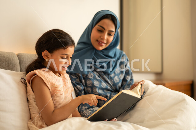 الاستمتاع بهواية مطالعة الكتب والروايات،  قضاء الوقت بالقراءه والتثقف، ثقافة التعلم الذاتي، قراءة القصص قبل النوم، امرأة عربية خليجية سعودية محجبة ترتدي العباءة تجلس على السرير مع ابنتها تحمل بيديها كتاب