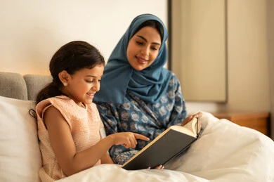 الاستمتاع بهواية مطالعة الكتب والروايات،  قضاء الوقت بالقراءه والتثقف، ثقافة التعلم الذاتي، قراءة القصص قبل النوم، امرأة عربية خليجية سعودية محجبة ترتدي العباءة تجلس على السرير مع ابنتها تحمل بيديها كتاب