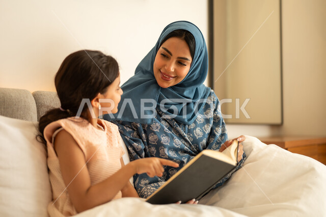 Spending time reading and educating oneself, the culture of self-learning, reading bedtime stories, enjoying the hobby of reading books and novels, an Arab Gulf Saudi woman wearing a hijab and an abaya sitting on the bed with her daughter holding a book in her hands.