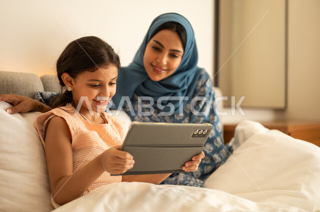 حضور الدروس الدراسية من خلال الإنترنت، المناهج العلمية التعليمية، فتاة عربية خليجية سعودية ترتدي الفستان تحمل بيدها الجهاز اللوحي، استخدام جهاز تقني حديث ومتطور، ام ترتدي العباءة تجلس مع ابتنها على السرير، مفهوم التعليم الالكتروني