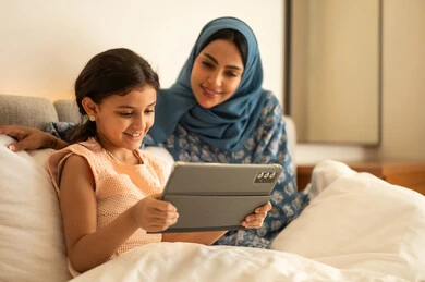 حضور الدروس الدراسية من خلال الإنترنت، المناهج العلمية التعليمية، فتاة عربية خليجية سعودية ترتدي الفستان تحمل بيدها الجهاز اللوحي، استخدام جهاز تقني حديث ومتطور، ام ترتدي العباءة تجلس مع ابتنها على السرير، مفهوم التعليم الالكتروني