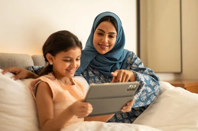 Following lessons remotely via the internet, educational scientific curricula, an Arab Gulf Saudi girl wearing a dress holding a tablet in her hand, using a modern and advanced technological device, a mother wearing an abaya sitting with her daughter on the bed, the concept of e-learning.