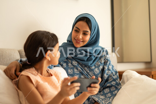 فتاة عربية خليجية سعودية ترتدي الفستان تحمل بيدها الهاتف المحمول، استخدام جهاز تقني حديث ومتطور، ام ترتدي العباءة تجلس مع ابتنها على السرير، مفهوم التعليم الالكتروني، متابعة الدروس عن بعد عن طريق الانترنت، المناهج العلمية التعليمية