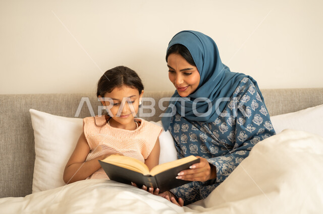 ثقافة التعلم الذاتي، قراءة القصص قبل النوم، قضاء الوقت بالقراءه والتثقف، الاستمتاع بهواية مطالعة الكتب والروايات، امرأة عربية خليجية سعودية محجبة ترتدي العباءة تجلس على السرير مع ابنتها تحمل بيديها كتاب