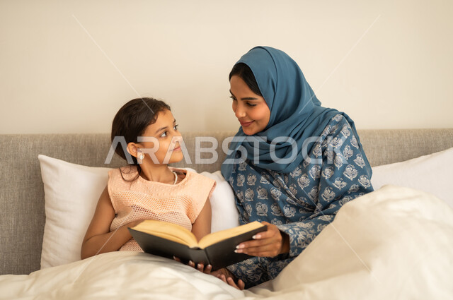 قراءة القصص قبل النوم، ثقافة التعلم الذاتي، قضاء الوقت بالقراءه والتثقف، الاستمتاع بهواية مطالعة الكتب والروايات، امرأة عربية خليجية سعودية محجبة ترتدي العباءة تجلس على السرير مع ابنتها تحمل بيديها كتاب