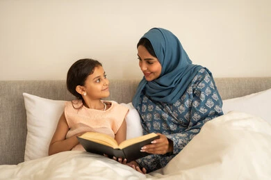 قراءة القصص قبل النوم، ثقافة التعلم الذاتي، قضاء الوقت بالقراءه والتثقف، الاستمتاع بهواية مطالعة الكتب والروايات، امرأة عربية خليجية سعودية محجبة ترتدي العباءة تجلس على السرير مع ابنتها تحمل بيديها كتاب