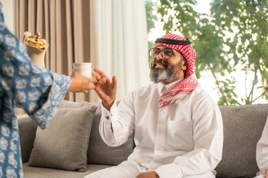 تقديم المشروبات الساخنة العربية الأصيلة في الأعياد، كرم الضيافة وحسن الاستقبال في السعودية، صورة مقربة ليد إمرأة عربية خليجية سعودية ترتدي العباءة تقوم بصب القهوة الخليجية وتقديمها للضيوف في صالة المنزل، أصالة عادات وتقاليد المجتمع السعودي في المناسبات الاسلامية