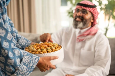 Facial expressions indicating happiness, a family gathering of a Saudi Arabian Gulf Arab family in the living room celebrating Eid, warm hospitality and honoring guests, a close-up image of the hand of a Saudi Arabian Gulf woman presenting sweets on the occasion of the happy Eid, joy and delight among family and relatives.