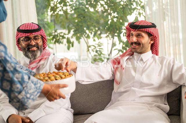 تجمع أسري لعائلة عربية خليجية سعودية في غرفة الجلوس يحتفلون في العيد، حسن الاستقبال واكرام الضيوف, صورة مقربة ليد امراه عربية خليجية سعودية تقوم بتقديم الحلوي في مناسبة العيد السعيد, البهجة والفرحة بين الاهل والاقارب, ايماءات وجه تدل على السعادة