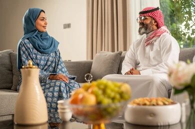 The ties of kinship and family bonding, gatherings and family visits during Islamic holidays and occasions, an Arab Gulf Saudi man wearing traditional attire sitting with his daughter with gestures of joy and happiness, hospitality and holiday sweets, good reception and honoring the guest.