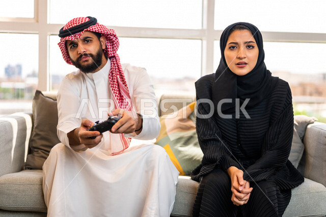 زوجان عربيان خليجيان سعوديان يجلسان في غرفة المعيشة ، قضاء وقت ممتع بلعب ألعاب الفيديو بإيماءات تدل على التركيز ، رجل يمسك عصا التحكم و يلعب ألعاب الفيديو بجانب زوجته ، المتعة و التسلية  بألعاب البلايستيشن الترفيهية ، مفهوم الأسرة السعيدة
