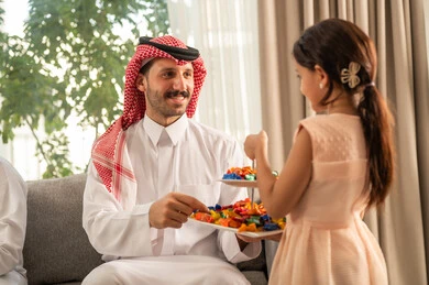 تقديم ضيافة وحلوى العيد، فرحة وبهجة الابناء في ايام العيد السعيد، حسن استقبال وإكرام الضيف، صلة الرحم والترابط الاسري, التجمعات والزيارات الاسرية في الأعياد والمناسبات الاسلامية, رجل عربي خليجي سعودي يرتدي الثوب التقليدي يجلس مع ابنته بإيماءات الفرح والسرور