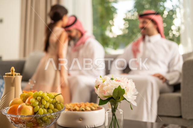 A Saudi Arabian Gulf man wearing traditional attire sits with his grandchildren, expressing joy and happiness. Family gatherings and visits during holidays and Islamic occasions, the joy and delight of children during the happy Eid days, hospitality and Eid sweets, good reception and honoring the guest, maintaining family ties and connections. A Saudi Arabian Gulf girl in a dress kisses her grandfather's head.