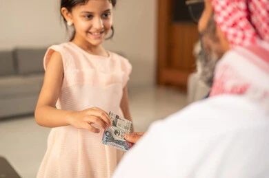 The role of the father in the lives of children, the concept of love and affection, giving Eid gifts during Eid, an Arab Gulf Saudi grandfather wearing traditional attire and a headscarf giving money to his granddaughter, customs and traditions of holidays and religious occasions, gestures of happiness and joy, family atmosphere filled with joy.