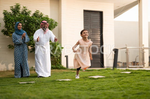 Caring for and nurturing children, maintaining family bonds, playing and having fun with the family outdoors, spending enjoyable time with children: a Saudi Arabian Gulf couple wearing traditional dress play in the park with their daughter, beaming with happiness.