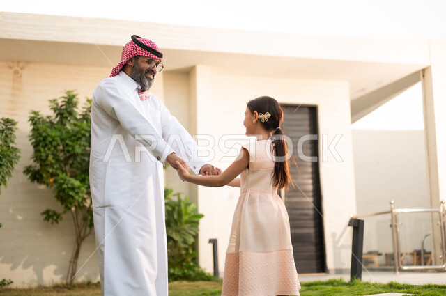 الترفيه والتسلية العائلية في الهواء الطلق, قضاء أوقات ممتعه مع الأبناء، رجل عربي خليجي سعودي يرتدي الثوب التقليدي والشماغ يلعب في الحديقة مع ابنته بإيماءات السعادة، المحافظة على الترابط الأسري، الاعتناء والاهتمام بالاولاد