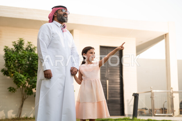 الاشارة الى شيء ما, قضاء أوقات ممتعه مع الأبناء، رجل عربي خليجي سعودي يرتدي الثوب التقليدي والشماغ يلعب في الحديقة مع ابنته بإيماءات السعادة، المحافظة على الترابط الأسري، الاعتناء والاهتمام بالاولاد