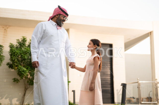 Enjoying recreational activities and playing with the family outdoors, spending enjoyable times with the children, an Arab Gulf Saudi man wearing traditional attire and a ghutrah playing in the park with his daughter with gestures of happiness, maintaining family bonds, caring for and paying attention to the children.