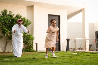 Playing and having fun with the family in outdoor settings, spending enjoyable times with the children, an Arab Gulf Saudi man wearing traditional attire and a headscarf playing in the garden with his daughter with gestures of happiness, maintaining family bonds, caring for and paying attention to the children.