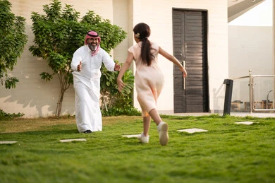 قضاء أوقات من اللعب والترفيه العائلي في الهواء الطلق, قضاء أوقات ممتعه مع الأبناء، رجل عربي خليجي سعودي يرتدي الثوب التقليدي والشماغ يلعب في الحديقة مع ابنته بإيماءات السعادة، المحافظة على الترابط الأسري، الاعتناء والاهتمام بالاولاد