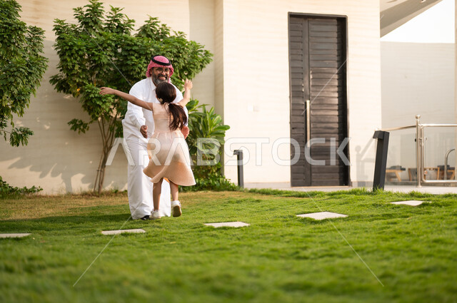 Enjoying playing, entertainment, and fun with the family outdoors, spending enjoyable times with the children, an Arab Gulf Saudi man wearing traditional attire and a shemagh playing in the garden with his daughter with gestures of happiness, maintaining family bonds, caring for and paying attention to the children.