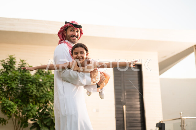 اللعب والتسلية والترفيه مع العائلة في الهواء الطلق، المحافظة على الترابط الأسري، قضاء أوقات ممتعه مع الأبناء، رجل عربي خليجي سعودي يرتدي الثوب التقليدي والشماغ يرفع ابنته عاليا بإيماءات السعادة، الاعتناء والاهتمام بالاولاد