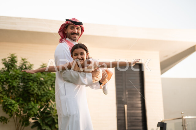 المحافظة على الترابط الأسري، قضاء أوقات ممتعه مع الأبناء، رجل عربي خليجي سعودي يرتدي الثوب التقليدي والشماغ يرفع ابنته عاليا بإيماءات السعادة، الاعتناء والاهتمام بالاولاد، اللعب والتسلية والترفيه مع العائلة في الهواء الطلق