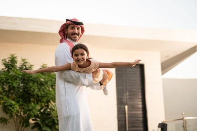 المحافظة على الترابط الأسري، قضاء أوقات ممتعه مع الأبناء، رجل عربي خليجي سعودي يرتدي الثوب التقليدي والشماغ يرفع ابنته عاليا بإيماءات السعادة، الاعتناء والاهتمام بالاولاد، اللعب والتسلية والترفيه مع العائلة في الهواء الطلق