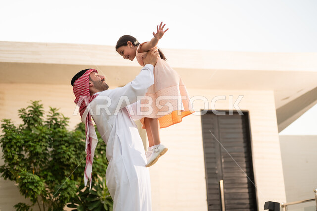 قضاء أوقات ممتعه مع الأبناء، رجل عربي خليجي سعودي يرتدي الثوب التقليدي والشماغ يرفع ابنته عاليا بإيماءات السعادة، المحافظة على الترابط الأسري، الاعتناء والاهتمام بالاولاد، اللعب والتسلية والترفيه مع العائلة في الهواء الطلق