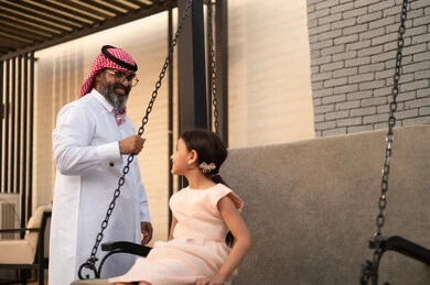 Spending a good time outdoors, enjoying playing on a sunny day, swinging on the swing, a smiling Arab Saudi Gulf girl playing in the park with expressions of joy and happiness alongside her father.