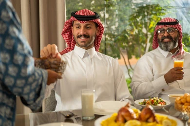 كسر الصيام بالتمر عند أذان المغرب في رمضان المبارك، فطور رمضاني عائلي، امراه عربيه خليجيه سعودية محجبة تقوم بتقديم الطعام, حسن الضيافة وإكرام الأخرين, أجواء عائلية روحانية دينية إسلامية, اجتماع الأسرة حول مائدة الإفطار المليئة بأصناف المأكولات والمقبلات الشهية