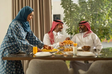 موعد كسر الصيام عند أذان المغرب في رمضان المبارك، فطور رمضاني عائلي، امراه عربيه خليجيه سعودية محجبة تقوم بتقديم الطعام على السفرة, حسن الضيافة وإكرام الأخرين, أجواء عائلية روحانية دينية إسلامية, اجتماع الأسرة حول مائدة الإفطار المليئة بأصناف المأكولات والمقبلات الشهية
