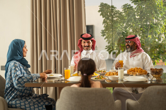 The family members gather around a table full of delicious dishes, a family iftar during Ramadan, the time to break the fast at the call to prayer at sunset in the blessed month of Ramadan. A Saudi Gulf Arab family sits at the table with gestures of joy, hospitality, and honoring others, creating a spiritual and religious Islamic family atmosphere.