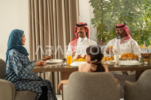 فطور رمضاني عائلي، اجتماع الأسرة حول مائدة الإفطار المليئة بأصناف المأكولات والمقبلات الشهية، موعد كسر الصيام عند أذان المغرب في رمضان المبارك، عائلة عربية خليجية سعودية تجلس على السفرة بايماءات السرور, حسن الضيافة وإكرام الأخرين, أجواء عائلية روحانية دينية إسلامية