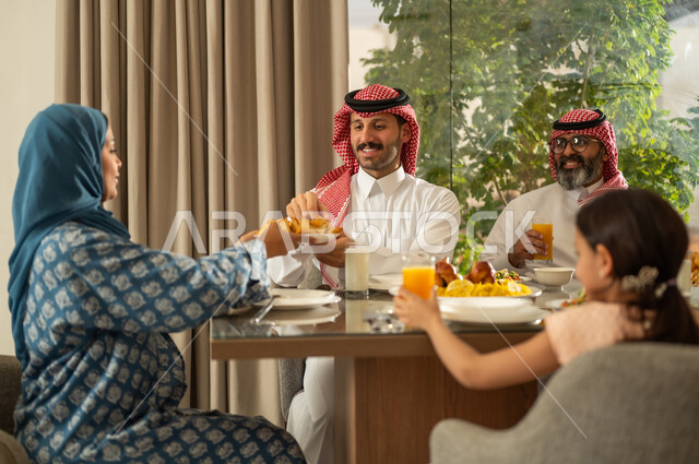 اجتماع الأسرة حول مائدة الإفطار المليئة بأصناف المأكولات والمقبلات الشهية، موعد كسر الصيام عند أذان المغرب في رمضان المبارك، فطور رمضاني عائلي، عائلة عربية خليجية سعودية تجلس على السفرة بايماءات السرور, حسن الضيافة وإكرام الأخرين, أجواء عائلية روحانية دينية إسلامية