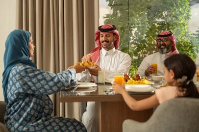The family gathers around the iftar table filled with a variety of delicious dishes and appetizers, the time to break the fast at the call to prayer at sunset during the blessed month of Ramadan, a family Ramadan breakfast, a Saudi Gulf Arab family sitting at the table with gestures of joy, hospitality, and honoring others, a spiritual and religious Islamic family atmosphere.