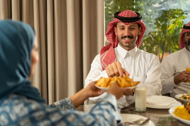 أجواء عائلية روحانية دينية إسلامية، حسن الضيافة وإكرام الأخرين, اجتماع الأسرة حول مائدة الإفطار المليئة بأصناف المأكولات والمقبلات الشهية, موعد كسر الصيام عند أذان المغرب في رمضان المبارك، فطور رمضاني عائلي، صورة مقرب لامراه عربيه خليجيه محجبة تقوم بتقديم طبق من السمبوسك