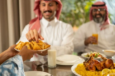 Hospitality and honoring others, a spiritual and religious Islamic family atmosphere, the family gathering around the iftar table filled with various delicious dishes and appetizers, the time to break the fast at the call to prayer at sunset during the blessed month of Ramadan, a family Ramadan breakfast, a close-up image of a Gulf Arab woman in a hijab serving a plate of samosas.