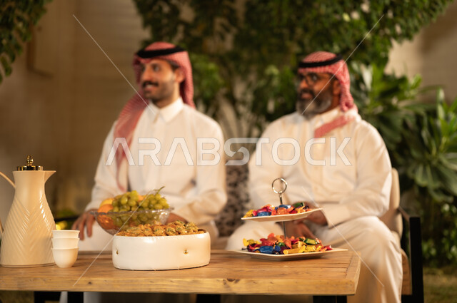 جلسة عائلية سعيدة، التسامر وتبادل الاحاديث، تعابير المودة والمحبة، الاستمتاع بقضاء وقت مع الابناء، رجل كبير بالسن عربي خليجي سعودي يرتدي الثوب التقليدي والشماغ يجلس  برفقة ابنه يقومان بتبادل الاحاديث بايماءات السعادة و السرور