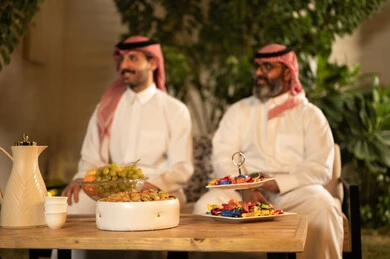 A happy family gathering, chatting and exchanging conversations, expressions of affection and love, enjoying time spent with the children. An elderly Arab Gulf Saudi man wearing traditional attire and a headscarf sits with his son, exchanging conversations with gestures of happiness and joy.