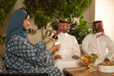 تقديم المشروبات الساخنة العربية الأصيلة في الأعياد، كرم الضيافة وحسن الاستقبال في السعودية، أصالة عادات وتقاليد المجتمع السعودي في المناسبات الاسلامية, إمرأة عربية خليجية سعودية ترتدي العباءة والحجاب تقوم بصب القهوة الخليجية وتقديمها للضيوف في صالة المنزل