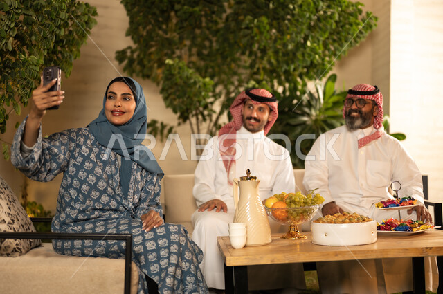 التقاط صورة سيلفي عبر الهاتف المحمول، ضيافة وحلوى العيد، حسن استقبال وإكرام الضيف، فرحة وبهجة الابناء في ايام العيد السعيد, التجمعات والزيارات الاسرية في الأعياد والمناسبات الاسلامية, رجل عربي خليجي سعودي يرتدي الثوب التقليدي يجلس مع أولاده بإيماءات الفرح والسرور