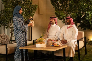 كرم الضيافة وحسن الاستقبال في السعودية، أصالة عادات وتقاليد المجتمع السعودي في المناسبات الاسلامية, إمرأة عربية خليجية سعودية ترتدي العباءة والحجاب تقوم بصب القهوة الخليجية وتقديمها للضيوف في صالة المنزل, تقديم المشروبات الساخنة العربية الأصيلة في الأعياد