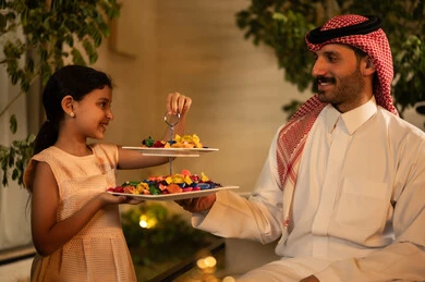 The warm welcome and honoring of guests, the ties of kinship and family bonding, the joy and happiness of children during the blessed Eid days, family gatherings and visits during Islamic holidays and occasions, an Arab Gulf Saudi man wearing traditional attire and a headscarf sitting with his daughter with gestures of joy and happiness, hospitality and Eid sweets.