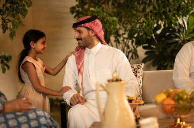حسن استقبال وإكرام الضيف، صلة الرحم والترابط الاسري, فرحة وبهجة الابناء في ايام العيد السعيد, التجمعات والزيارات الاسرية في الأعياد والمناسبات الاسلامية, رجل عربي خليجي سعودي يرتدي الثوب التقليدي يجلس مع ابنته تعابير الفرح والسرور، ضيافة وحلوى العيد