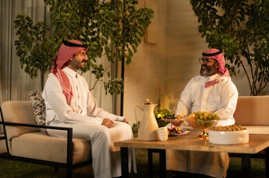 The atmosphere of the Saudi family during Eid, two Arab Gulf men from Saudi Arabia wearing the thobe and shemagh are exchanging conversation in a friendly environment, gestures of happiness and joy, offering Arabic hospitality with fruits and coffee, the concept of hospitality and social communication.