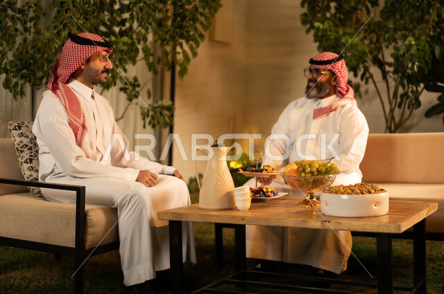 رجلان عربيان خليجيان سعوديان يرتديان الثوب والشماغ يقومان بتبادل الحديث في أجواء ودية، ايماءات السعادة و السرور، تقديم الضيافة العربية مع الفاكهة والقهوة، مفهوم الضيافة والتواصل الاجتماعي