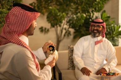 The atmosphere of the Saudi family during Eid, hospitality and reception, an Arab Gulf Saudi man wearing a thobe and shemagh pouring and serving hot coffee, the joy and happiness of Eid among family and relatives, Eid traditions in Saudi Gulf society, family visits and gatherings during holidays and occasions.