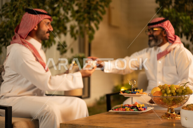 كرم الضيافة وحسن الاستقبال في السعودية، أصالة عادات وتقاليد المجتمع السعودي في المناسبات الاسلامية,رجل عربي خليجي سعودي يرتدي الثوب و الشماغ يقوم يتقديم القهوة الخليجية للضيف, تقديم المشروبات الساخنة العربية الأصيلة في الأعياد