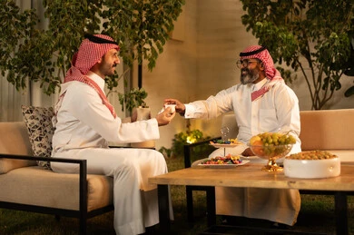 أصالة عادات وتقاليد المجتمع السعودي في المناسبات الاسلامية, كرم الضيافة وحسن الاستقبال في السعودية,رجل عربي خليجي سعودي يرتدي الثوب و الشماغ يقوم يتقديم القهوة الخليجية للضيف, تقديم المشروبات الساخنة العربية الأصيلة في الأعياد